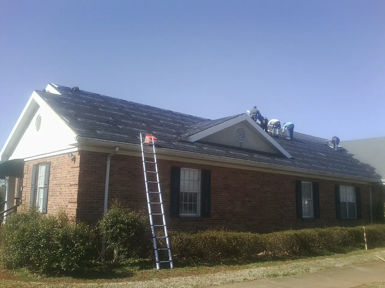 Crew tearing shingles off a brick ranch roof with a ladder set — Emergency Roof Repair work by our crew in Dudley